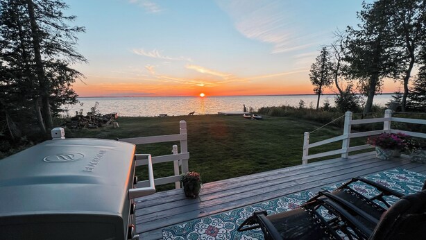 En terrass med utsikt över solnedgången över vattnet, en grill och ett vitt staket.