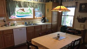 A kitchen with wooden cabinets, a white dishwasher, a table with a tablecloth, and a hanging lamp. - Felker's Cottage: Fall Color Getaway | Lakefront Cottage Near Tunnel of Trees (Vanderbilt)