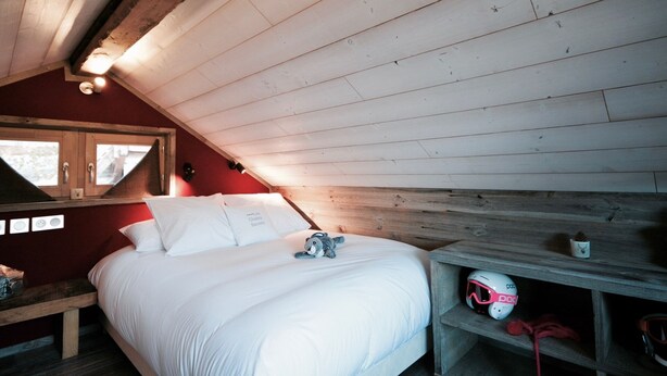 Quarto com teto de madeira, cama com roupa de cama branca, mesa de cabeceira com um capacete e janela com cortinas.