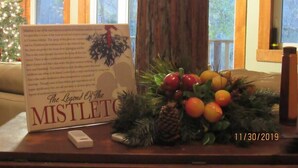 A table with a sign titled 'The Legend of the Mistletoe', a fruit arrangement, and a Christmas tree. - On the Riverbend Cabin (Claxton)