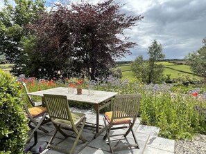 An outdoor dining area with a wooden table and chairs, surrounded by lush greenery and flowering plants.