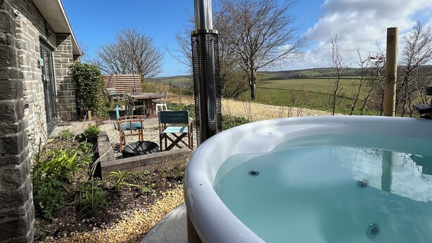 A hot tub in a backyard with a stone building, outdoor furniture, and a view of the countryside.