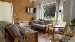 A modern living room with a wooden sofa, a grey armchair, and a wooden chair. There is a television mounted on the wall, a small table with a lamp, and a window with curtains.