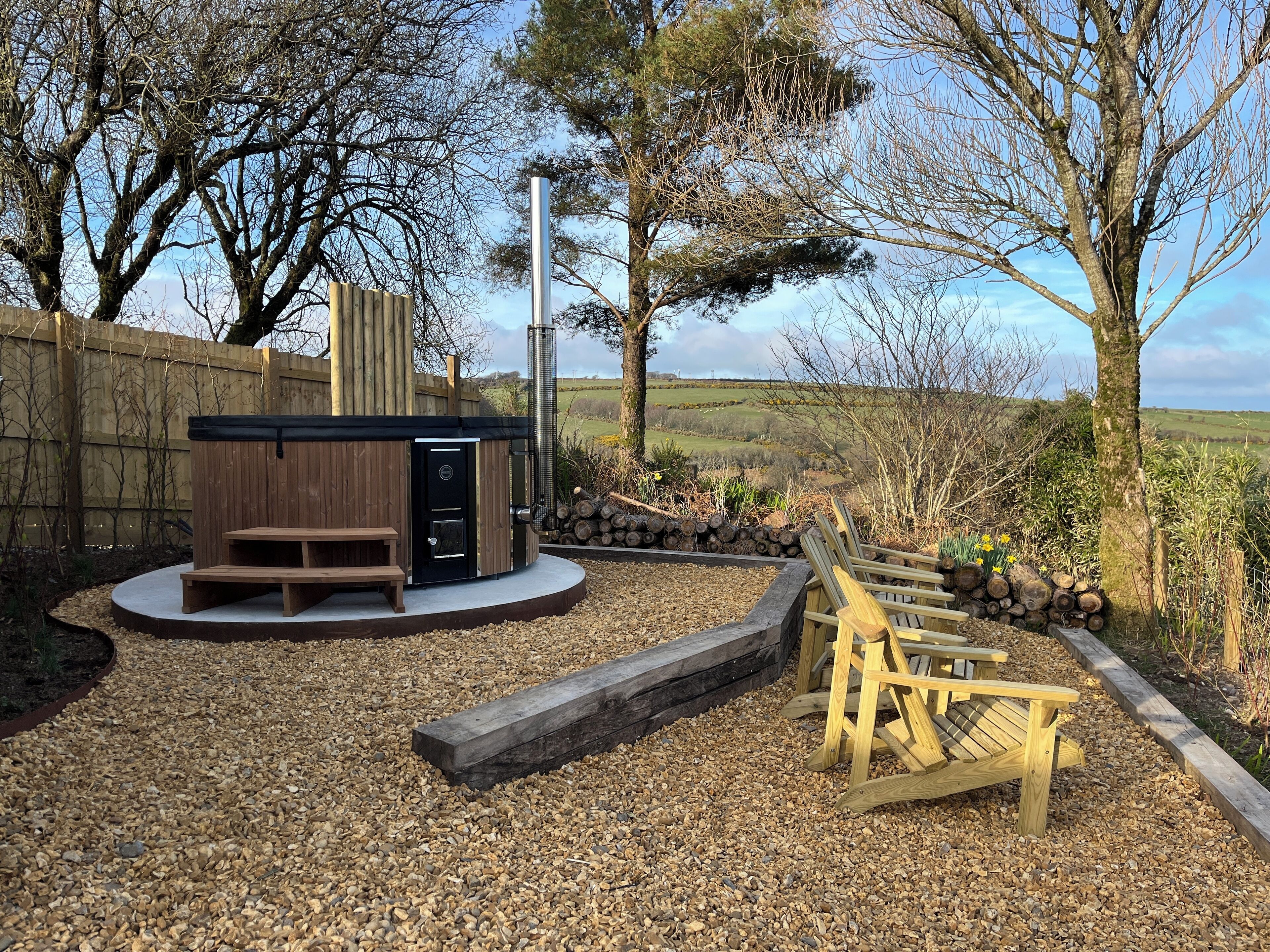 A hot tub with a wooden deck and a bench, surrounded by a gravel area and a wooden fence.