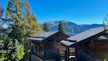 Uma cabana com telhado inclinado, rodeada por árvores, e vista para as montanhas ao fundo.