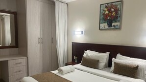 Un lit bien fait, avec des draps blancs et un plaid marron, une armoire en bois, une table de chevet avec une lampe, et un tableau de fleurs accroché au mur.