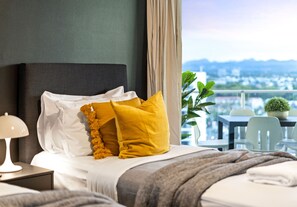 Ein Hotelzimmer mit Bett, Nachttisch, Lampe, Blick auf eine Stadtlandschaft durch große Fenster und einem Balkon mit Stühlen und einem Blumentopf.