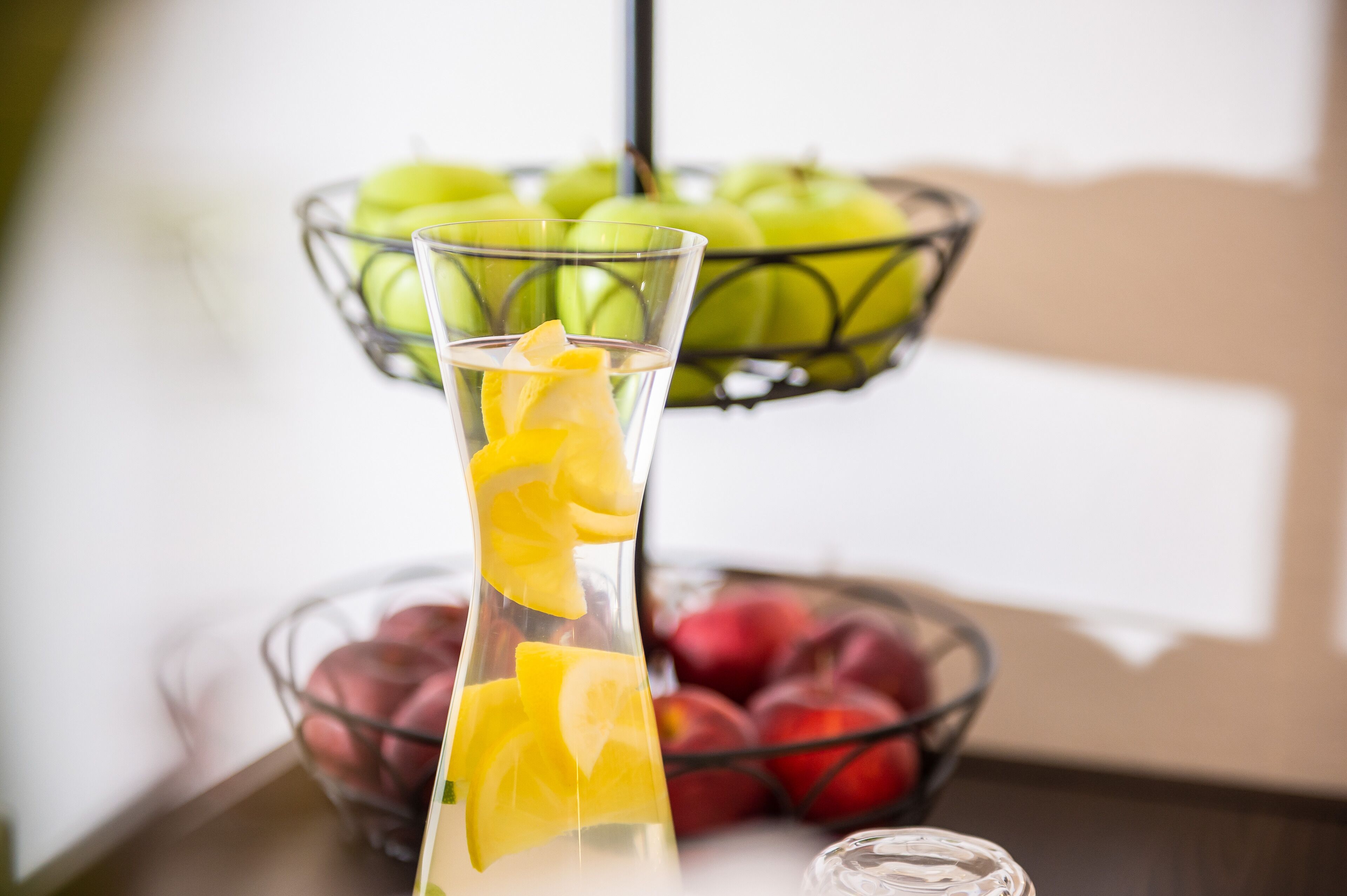 A clear glass vase with orange slices and a metal fruit basket containing green and red apples in the background.
