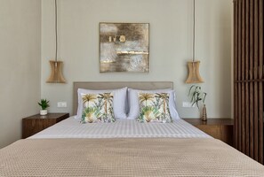 A neatly made bed with a striped bedspread, two decorative pillows with a tropical print, a wall-mounted abstract painting, and two wall-mounted lamps.