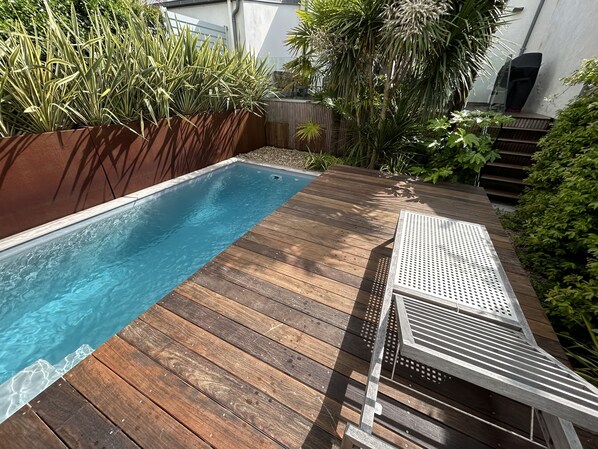 Une terrasse en bois avec une piscine, entourée de différentes plantes et agrémentée d’une chaise en métal.