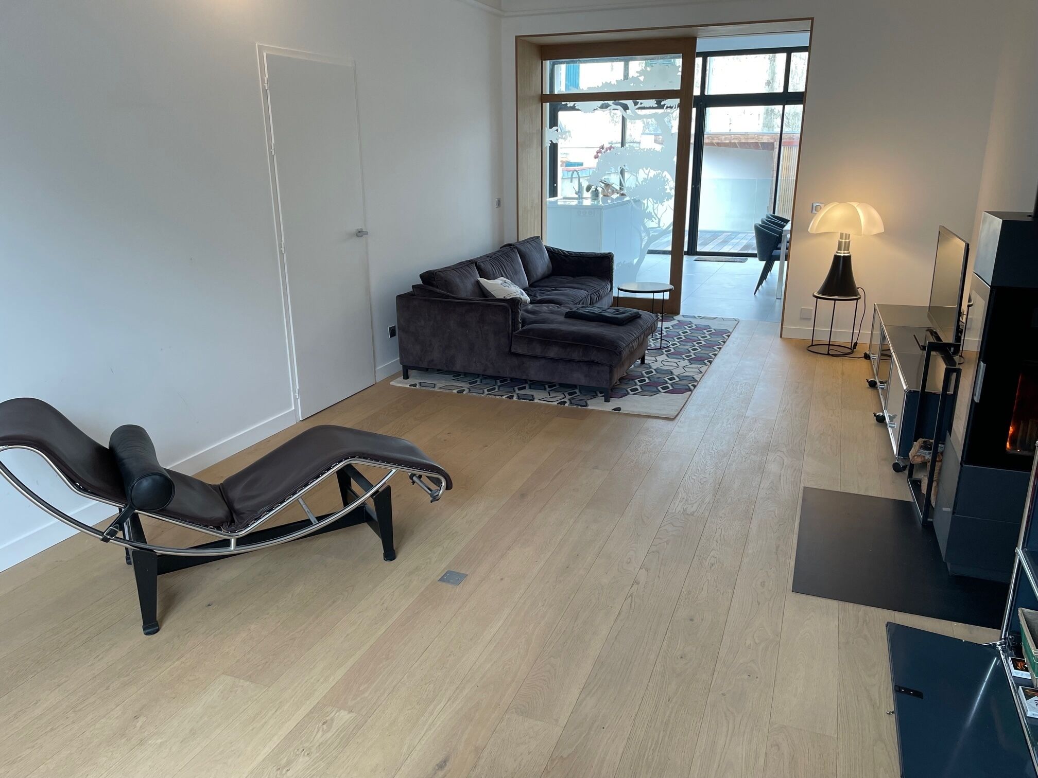 A modern living room with a sofa, a reclining chair, a small table, and a kitchen area with a stove and cabinets.