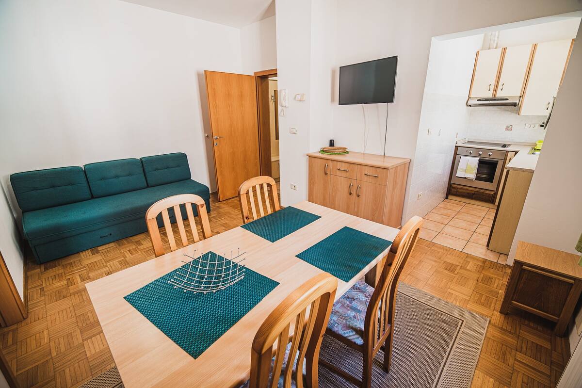 A compact living space with a wooden dining table, chairs, a green sofa, a TV, and a kitchenette.
