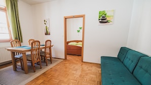 Ein Zimmer mit Holzboden, einem smaragdgrünen Sofa, einem Esstisch mit Stühlen, einem Bett mit grüner Tagesdecke und einem Bild an der Wand.
