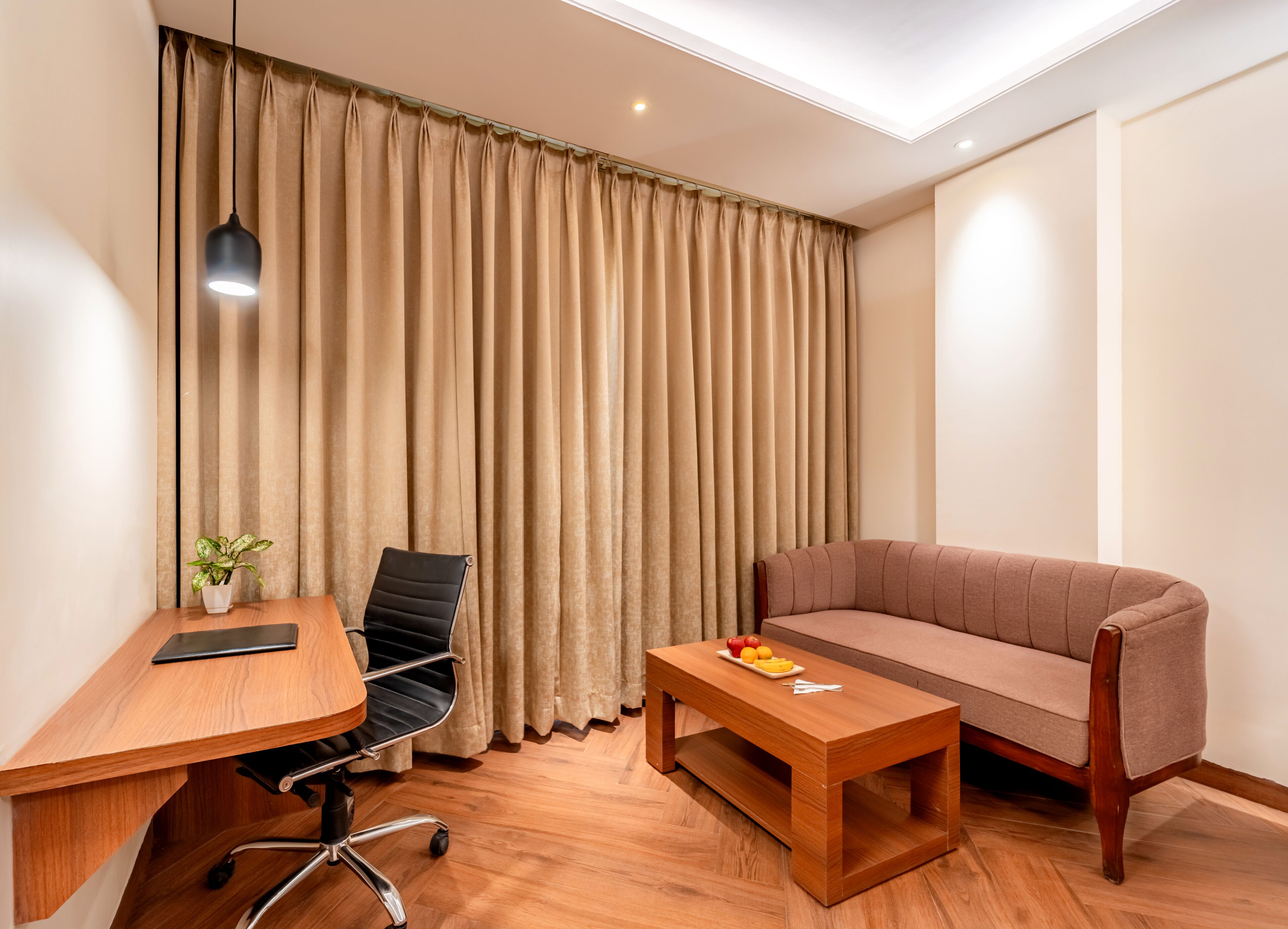A modern room with a wooden desk, a black office chair, a wooden coffee table, a beige sofa, and a large window with beige curtains.