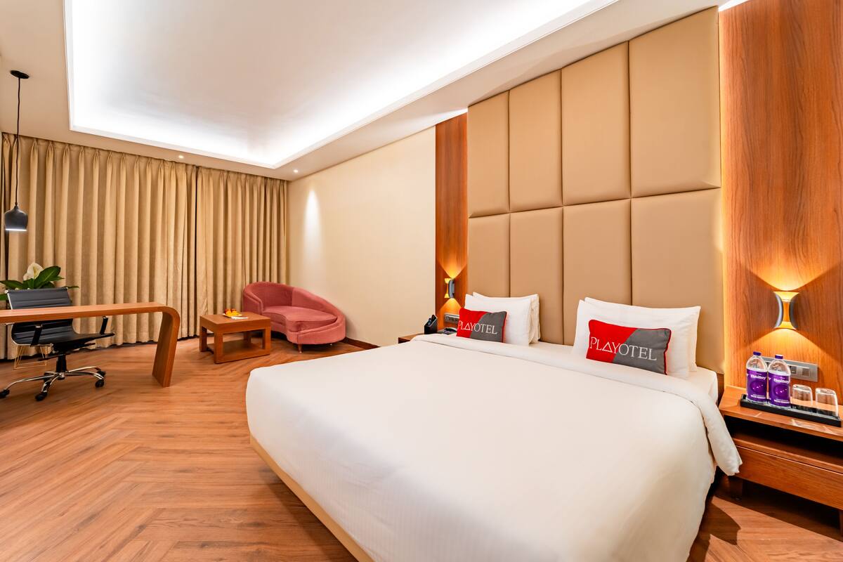 A hotel room with a large bed, a desk, a chair, and a bedside table with a lamp.