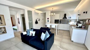 A modern living room with a dark blue sofa, a dining area with a white table and chairs, and a kitchen with white cabinets and a black range hood.