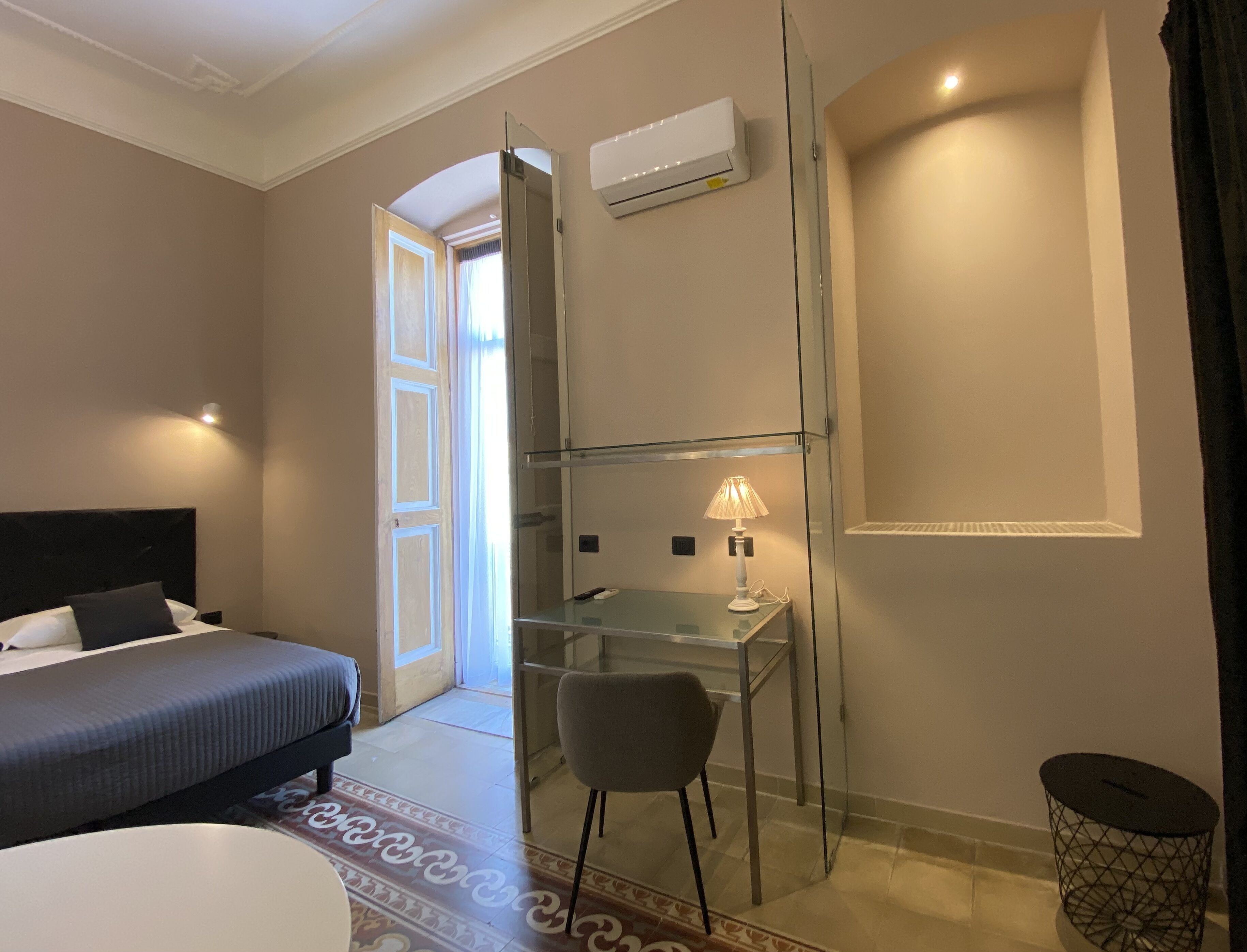 Habitación de hotel con cama, escritorio, silla, aire acondicionado y un cubo de basura pequeño.