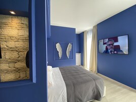 Un dormitorio con pared de piedra, una cama con colcha gris, dos adornos de pared tipo ala, y un televisor mostrando a una persona.