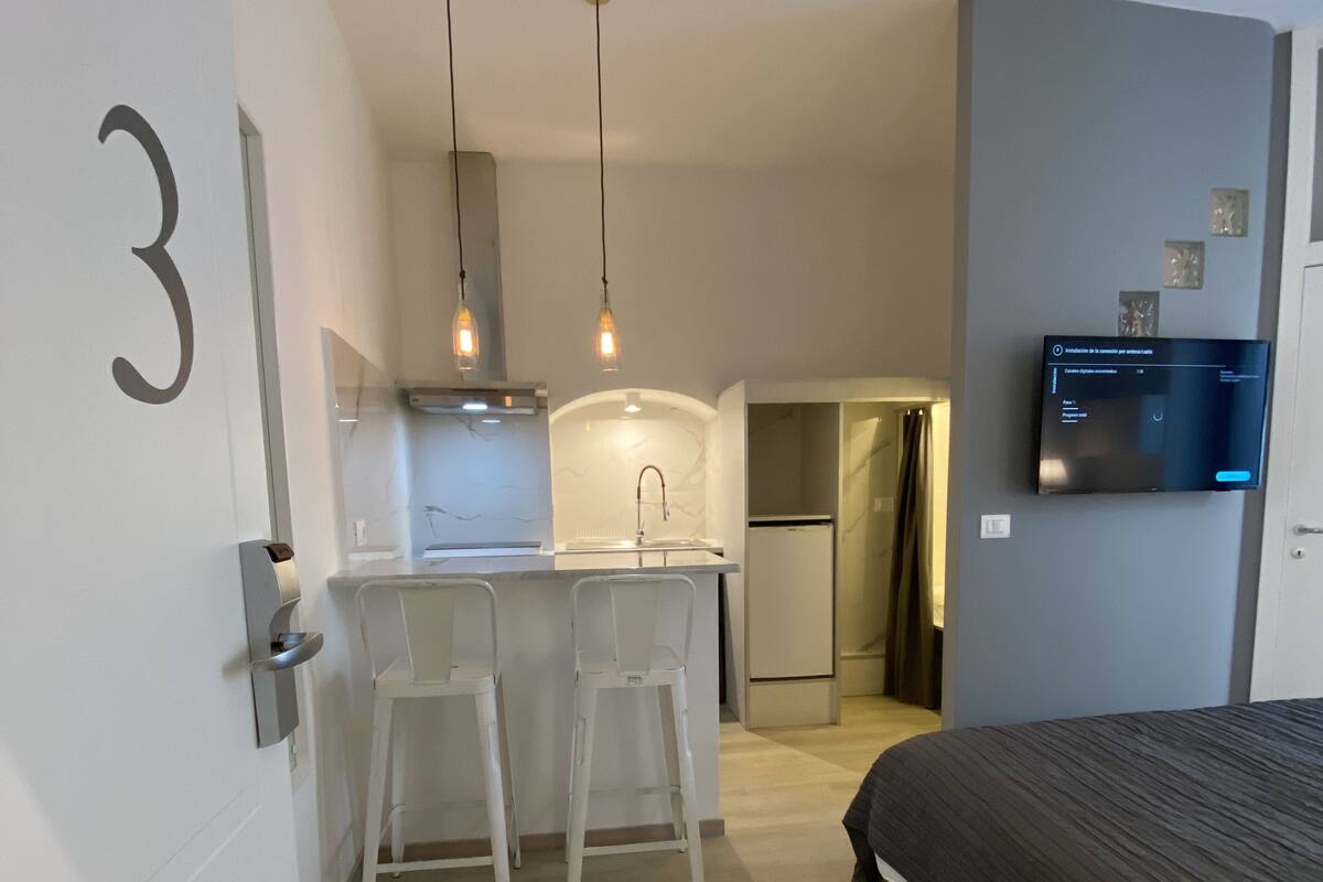 A modern bedroom with a large bed, a small kitchenette with a fridge, and a TV mounted on the wall.
