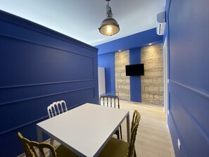 Quarto com mesa e cadeiras brancas, televisão na parede e um painel de pedra decorativa.
