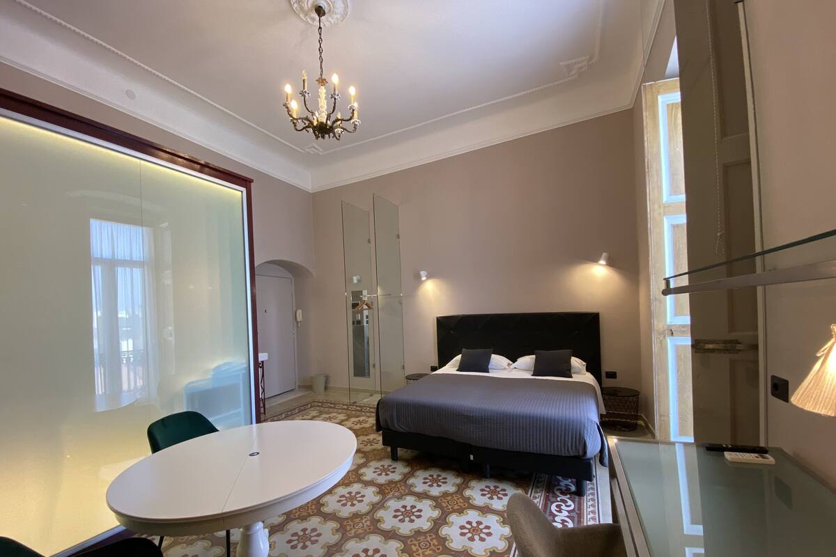 A hotel room with a bed, a glass table, a chair, a chandelier, and a mirror.