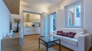Ein modernes Wohnzimmer mit einem Glastisch, einem weißen Sofa mit Kissen, einer Küche im Hintergrund und einer Treppe zu einer weiteren Ebene.