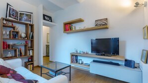Ein modernes Wohnzimmer mit einem weißen Sofa, einem Glastisch, einem Flachbildfernseher, Holzregalen mit Büchern und Dekoration sowie einer weißen Unterhaltungsecke.