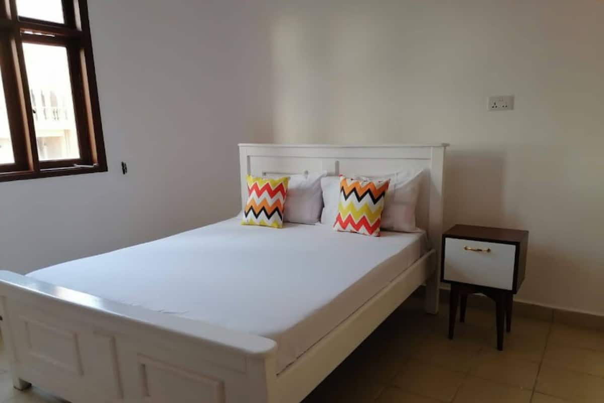 A single-bed room with a white bed, two colorful pillows, a small bedside table with a drawer, and a window with blinds.