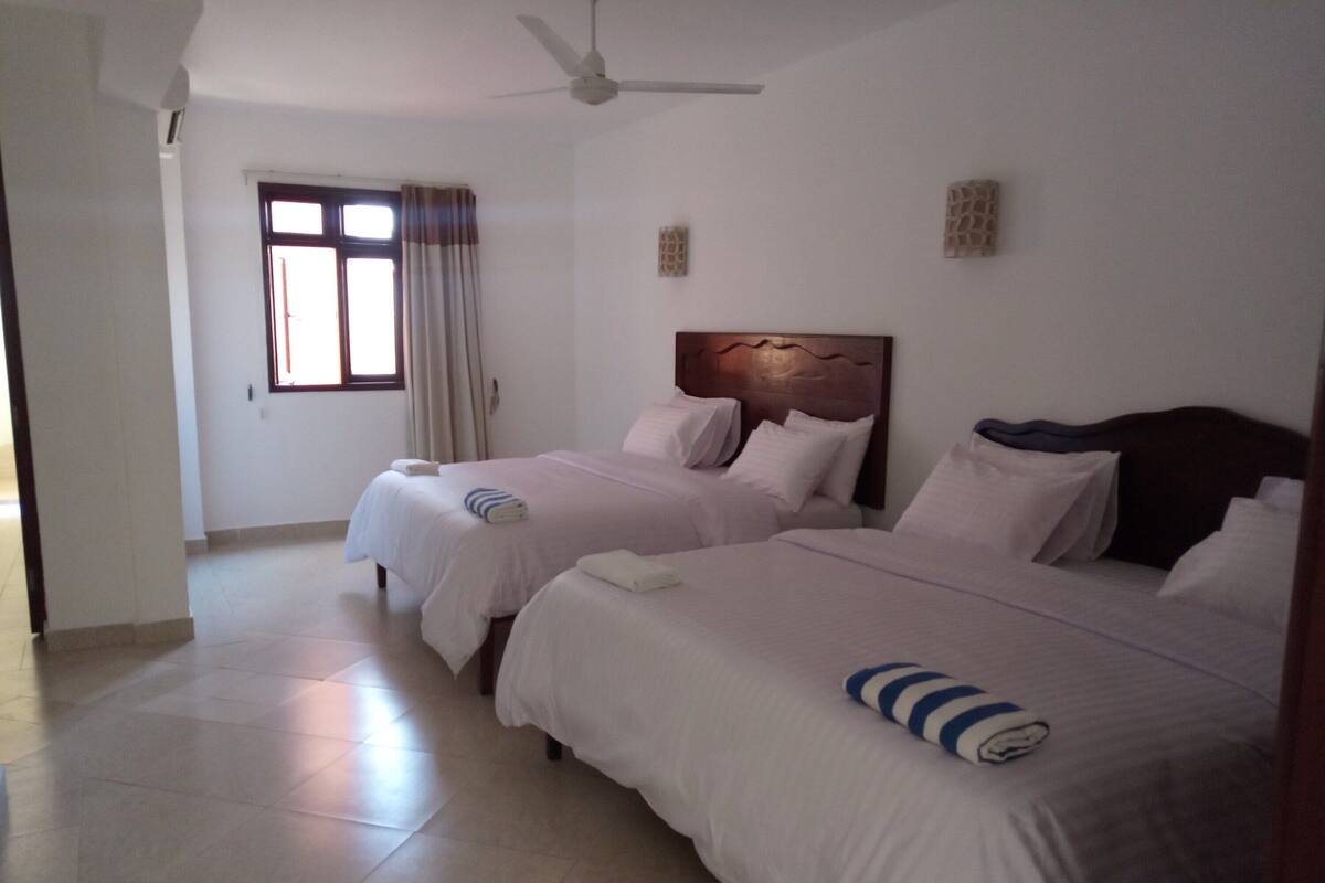 A hotel room with two beds, a ceiling fan, a window with curtains, and a tiled floor.