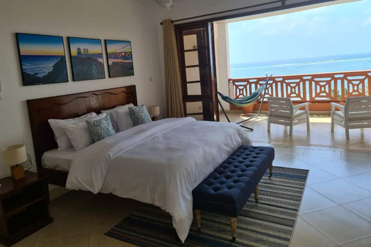 A bedroom with a bed, a nightstand, a bench, a hammock, and a balcony with a view of the ocean.