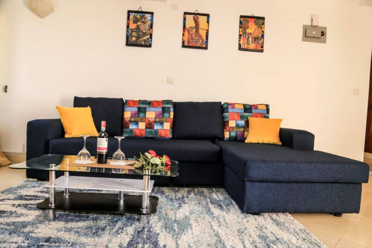 A modern living room with a dark blue sectional sofa, colorful cushions, a glass coffee table with wine glasses and a bottle, and a blue and white rug.