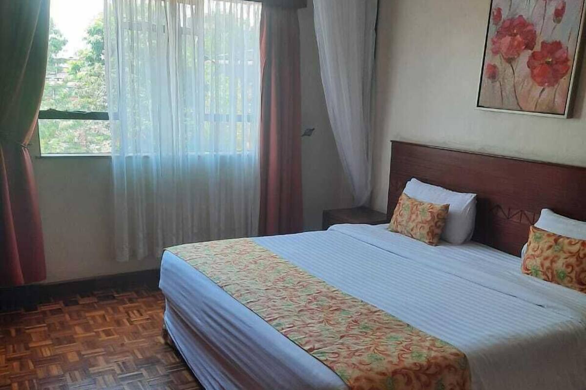 A hotel room with a large bed, wooden parquet flooring, a window with sheer curtains, and a floral painting on the wall.