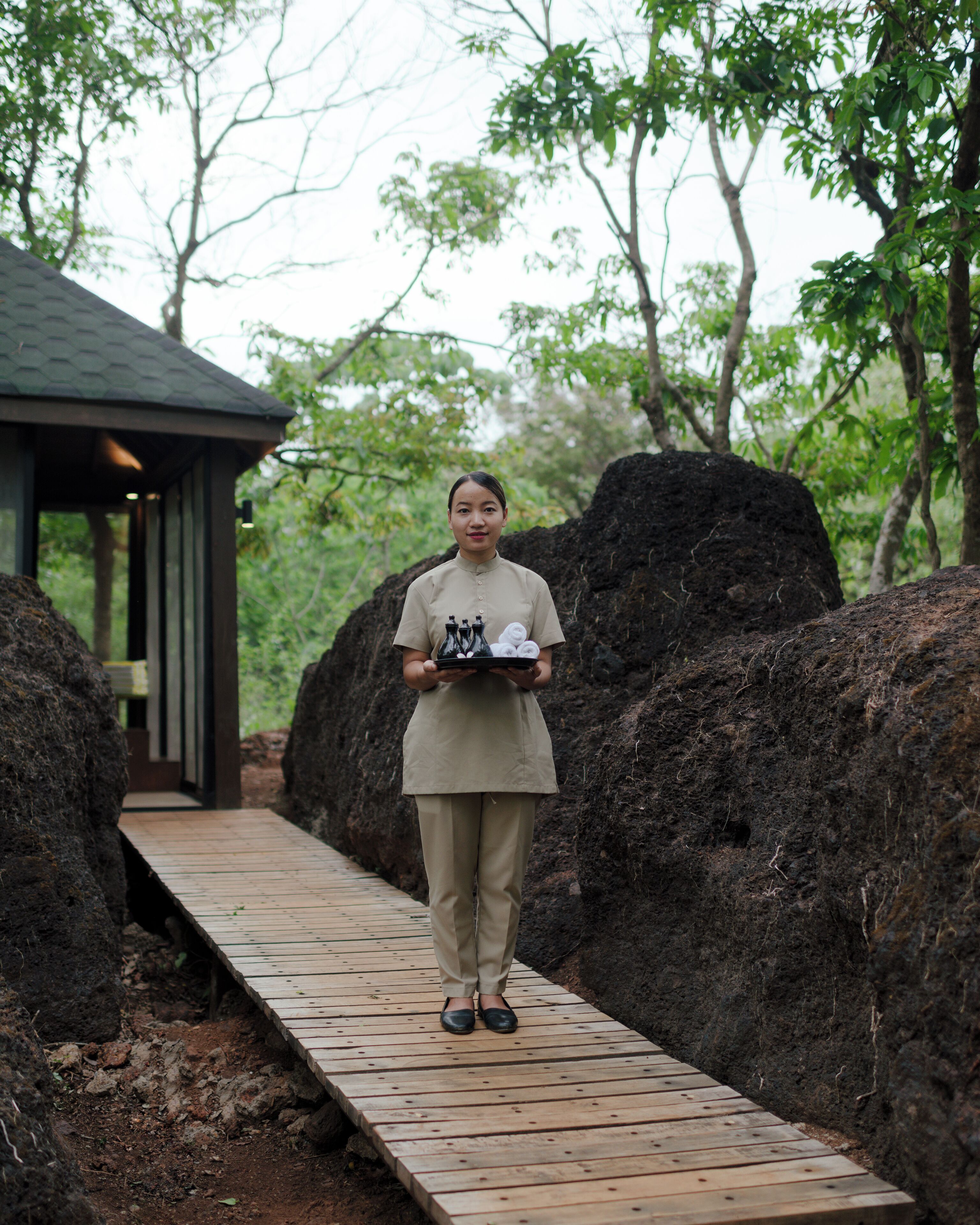 A person standing on a wooden pathway in a natural setting with large rocks on either side, leading to a building with a dark roof.
