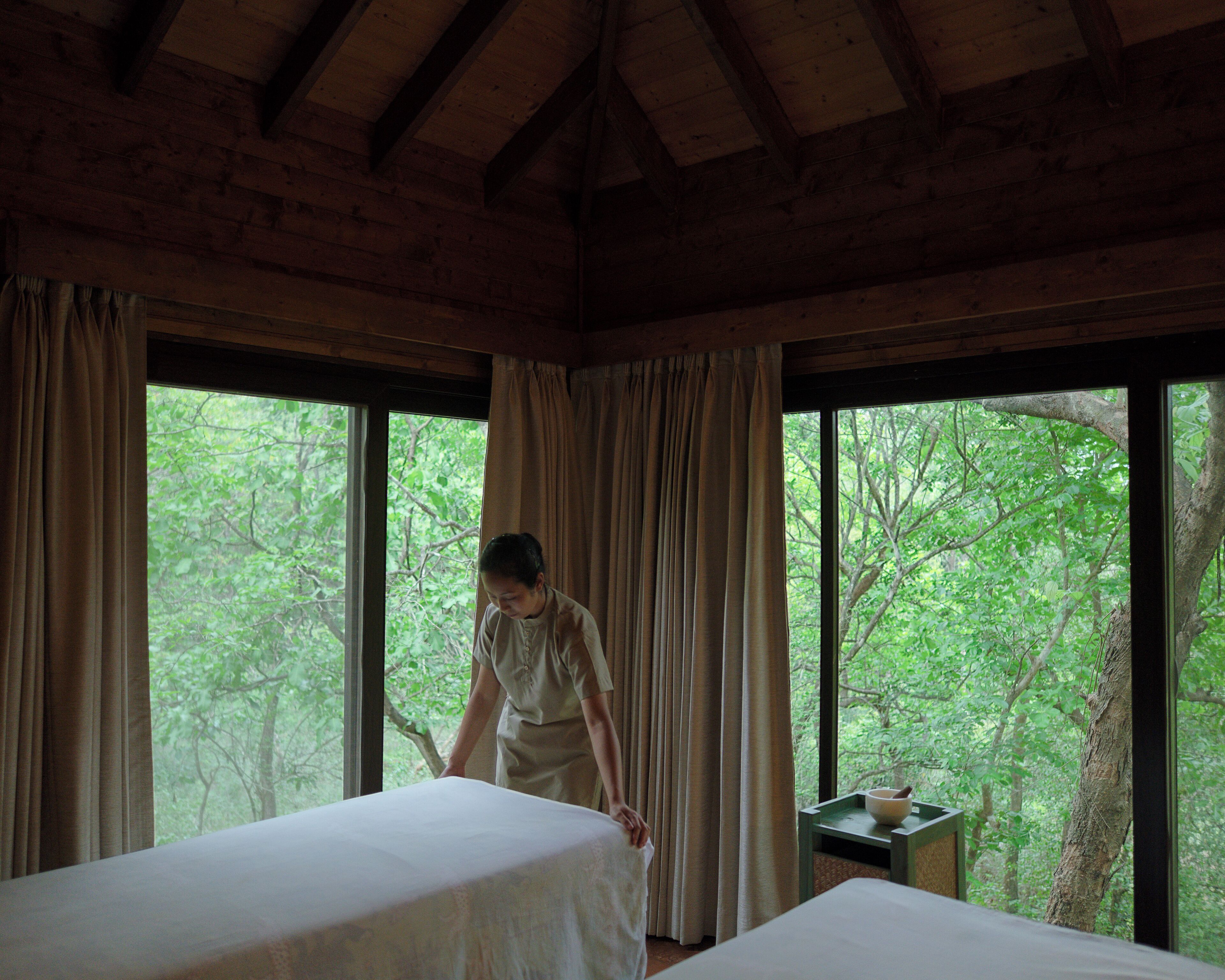 A room with two beds, large windows, and a view of trees.
