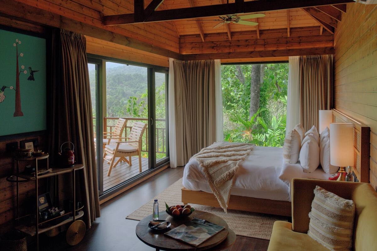 A bedroom with a large bed, a sitting area with a chair, a small table, and a view of the outdoors.