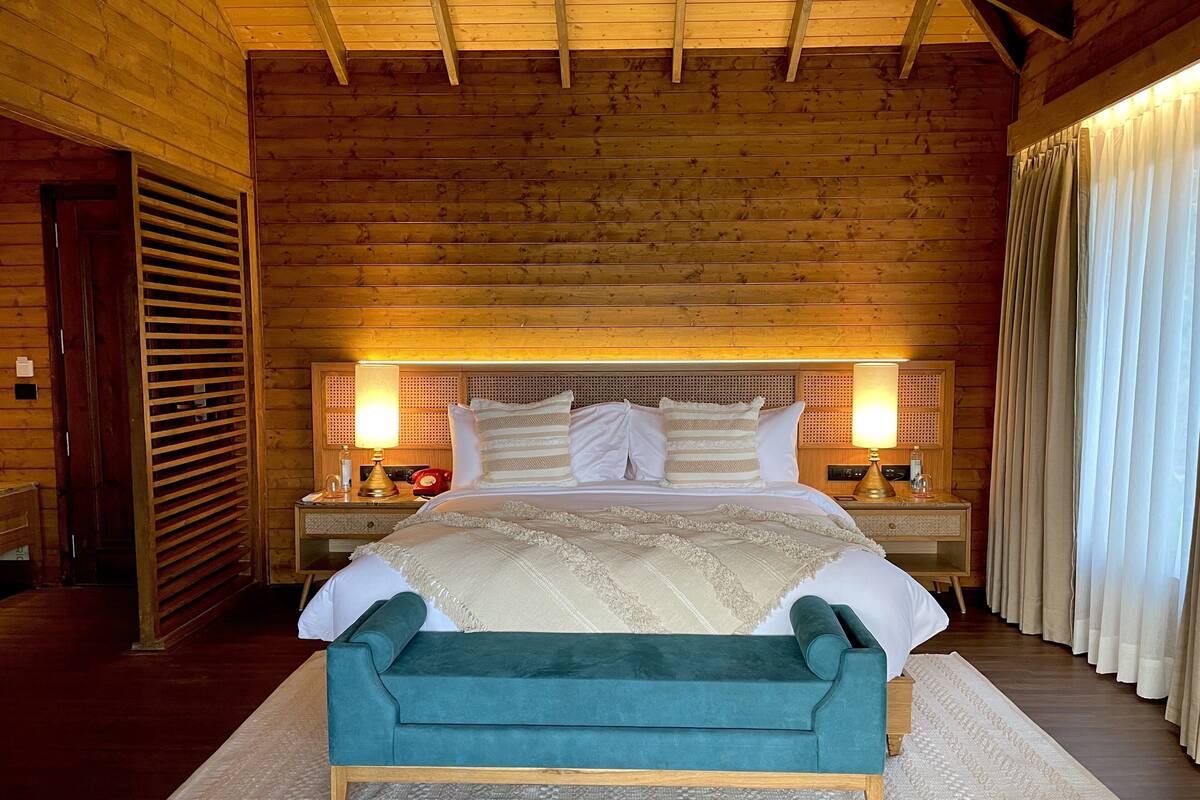 A wooden-paneled room with a bed, two bedside tables, a bench, and a window with curtains.