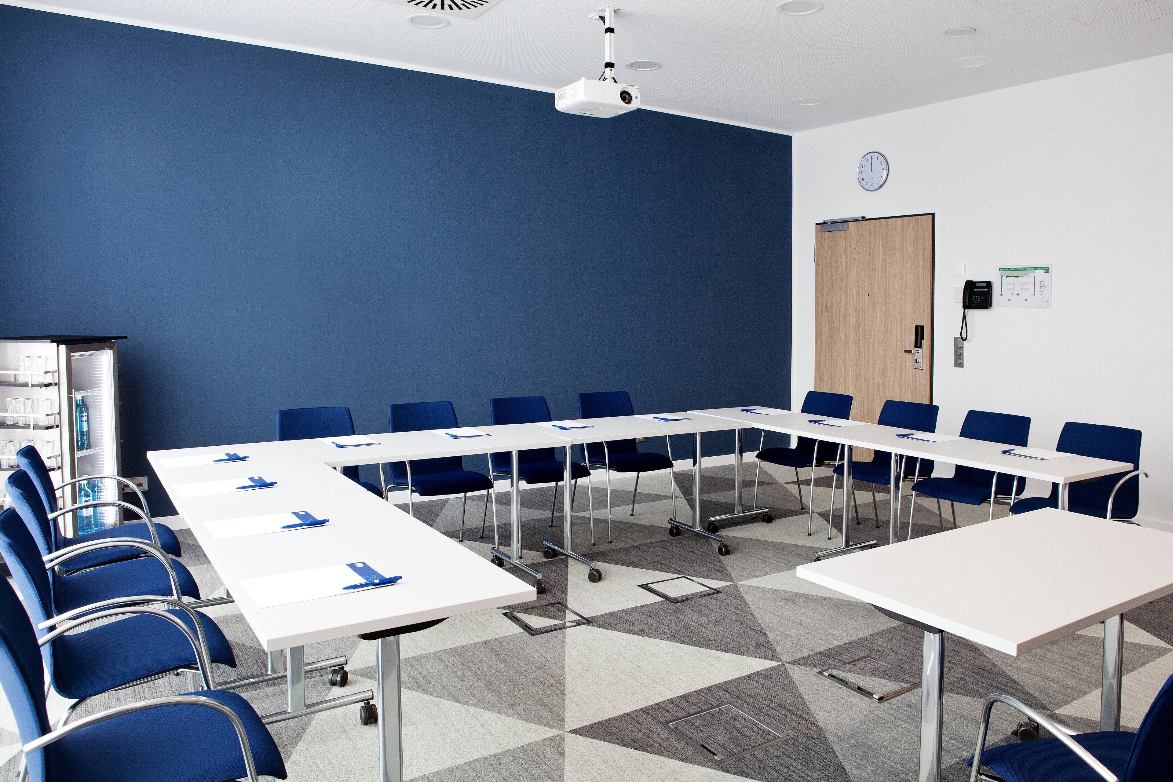 A conference room with a white table, blue chairs, a projector, and a clock on the wall.