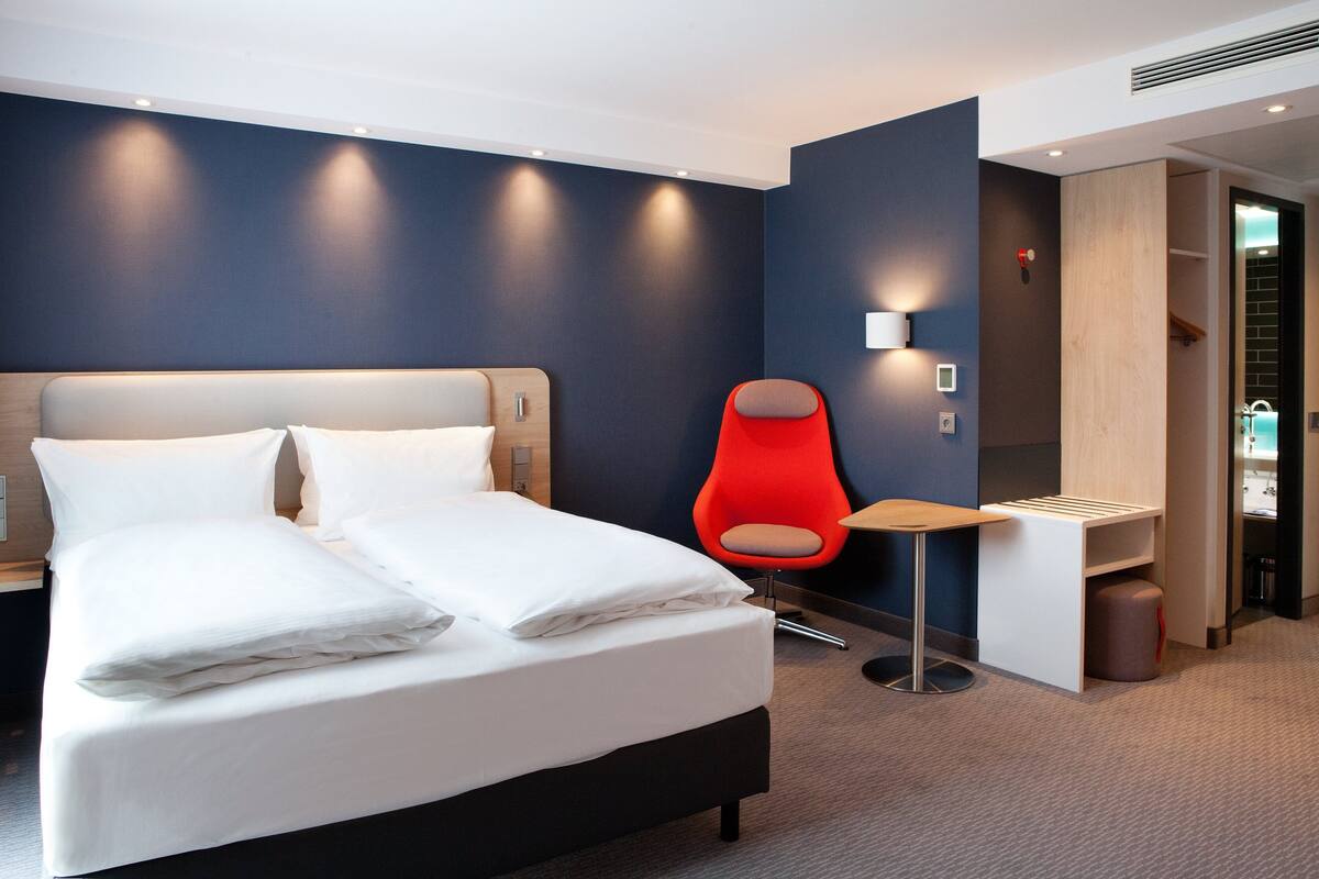 A modern hotel room with a large bed, a red chair, and a small table.