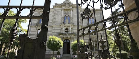 Una entrada imponente con rejas de metal ornamentales que conducen a un edificio histórico con un acceso central, flanqueado por árboles simétricos y un jardín bien cuidado.