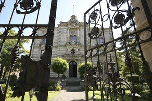 Una entrada imponente con rejas de metal ornamentales que conducen a un edificio histórico con un acceso central, flanqueado por árboles simétricos y un jardín bien cuidado.