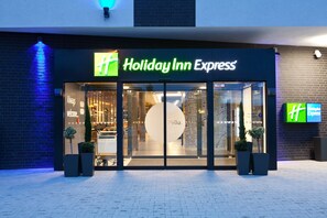 A Holiday Inn Express hotel entrance with a glass facade, a "Welcome" sign, and two potted plants in front.