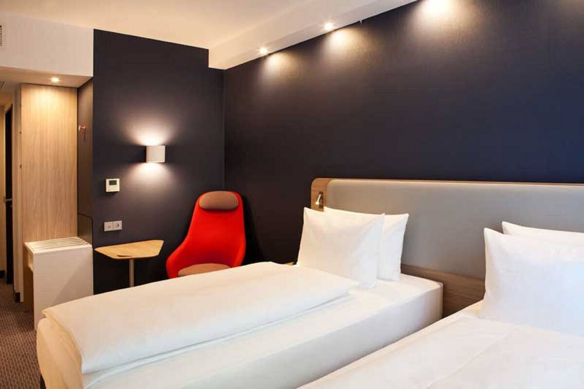 A hotel room with two beds, a red chair, a small table, and a wall-mounted light.