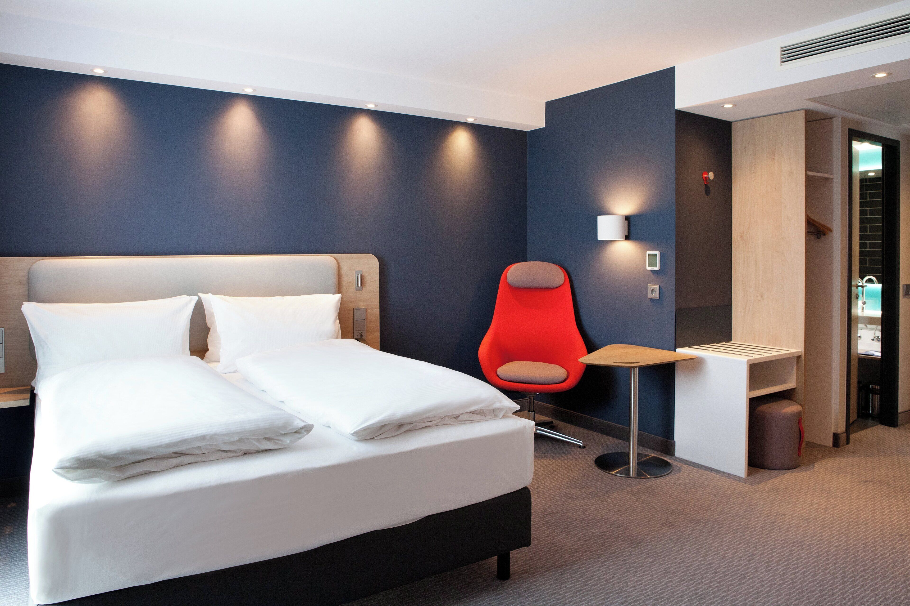 A hotel room with a bed, a red chair, a bedside table, and a wall-mounted light.