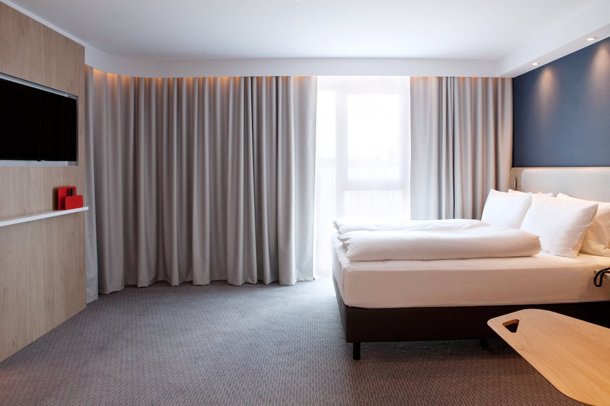 A modern hotel room with a large bed, a flat-screen TV mounted on the wall, and a wooden side table.