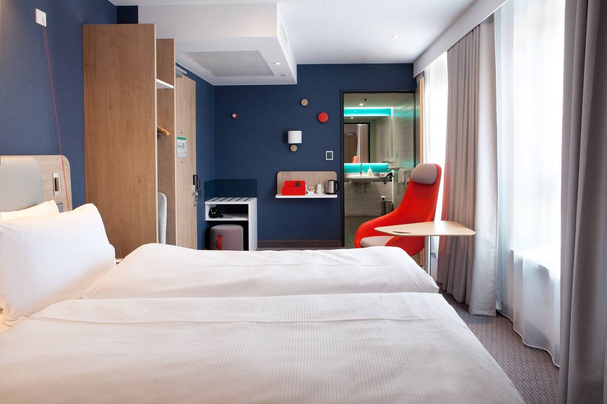 A hotel room with a large bed, a desk with a chair, a red chair, a small kitchenette, and a bathroom visible through a glass partition.