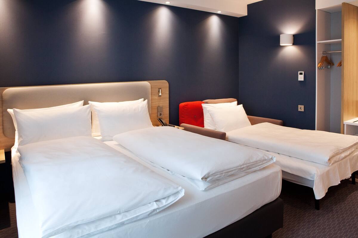 A hotel room with two beds, a wooden headboard, a red accent pillow, a bedside table, a mirror, and a wardrobe with hanging clothes.