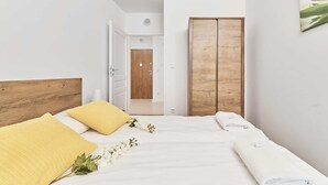 Un dormitorio con cabecera de madera, cama con ropa blanca y almohada amarilla, armario de madera y una puerta que da a otra habitación.