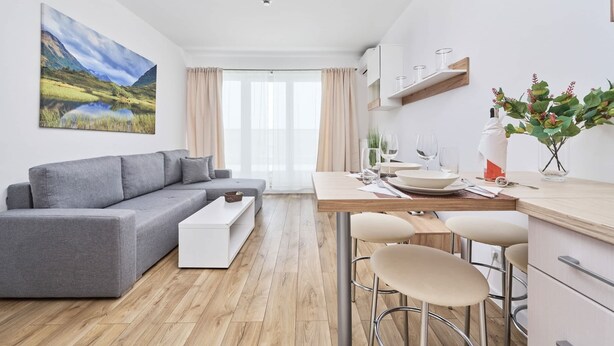 Una sala moderna con un sofá gris, un comedor con taburetes de bar, una cocina con armarios blancos y un cuadro de paisaje grande en la pared.