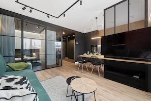 A modern living room with a sofa, a dining area with chairs, a TV, and a sliding glass door leading to a balcony. - Modern Apartment in Wroclaw | Balcony and Parking (Wroclaw)