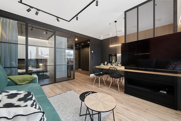 A modern living room with a sofa, a dining area with chairs, a TV, and a sliding glass door leading to a balcony.